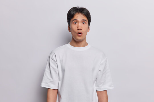 Waist up shot of stunned Asian man with dark hair stands impressed feels shocked dressed in casual basic t shirt poses against white background expresses surprisement. People and emotions concept