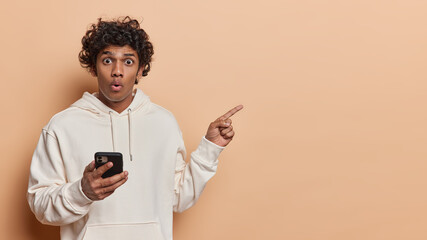 Waist up shot of surprised Hindu man texting to family sending sms browsing web points index finger...