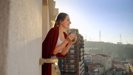 Beautiful woman standing on balcony with cup of coffee and looking at the city view in morning.