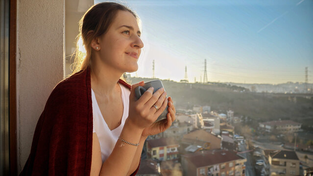 Portrait of smiling woman holding cup of tea and looking on city from balcony. People relaxing, resting at home, beautiful cityscape.