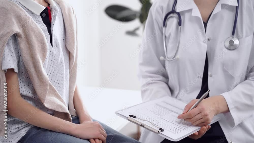 Wall mural Doctor and kid patient are in clinic, closeup. Unknown female physician using clipboard to fill out medical form near little boy. Medicine concept