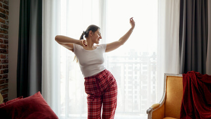 Happy smiling woman listening music and dancing in pajamas at window. People relaxing at home,...