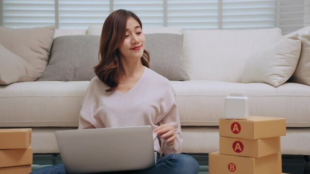 Young asian woman selling online receiving order via laptop and using label printer to printing customer address attach to the parcel box. Business Online and delivery shop concept