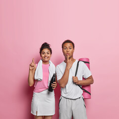 Pair of young fit happy smiling broadly European female and surprised African guy wearing sporty clothes preparing for competition keeping fingers crossed for good luck astonished to see opponent