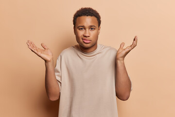 Studio shot of young confused African male with curly hair wearing casual tshirt isolated in centre on beige background shruggling shoulders spreading palms with hesitation. People emotions concept.