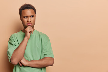 Emotions. Young thoughtful African male wearing casual green tshirt standing on left isolated on beige background trying to solve difficult problem holding one hand on chin with space for promotion