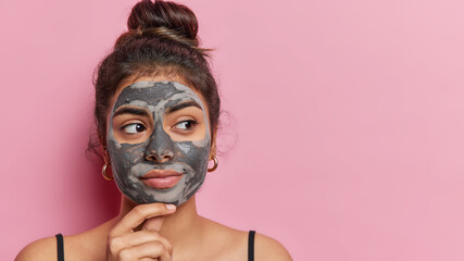 Beauty and wellness concept. Indoor portrait of european pretty young female applying natural beauty clay mask to keep her face fresh and clean isolated on pink background little bit enigmatic