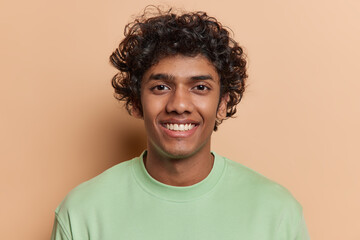 Portrait of handsome curly haired Hindu man smiles broadly has cheerful friendly expression white...
