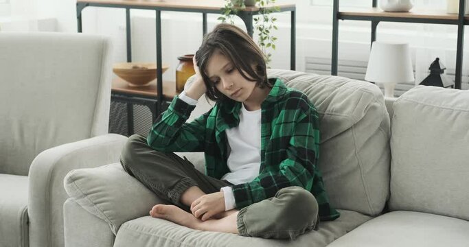 A young caucasian boy sits on the couch in the living room, his gaze fixed into the distance. His face reflects a profound sense of sadness and contemplation.