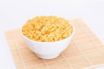 Uncooked Chifferi Rigati Pasta in White Cup on Bamboo Mat on White Background. Fat and Unhealthy Food. Scattered Classic Dry Macaroni. Italian Culture and Cuisine. Raw Pasta