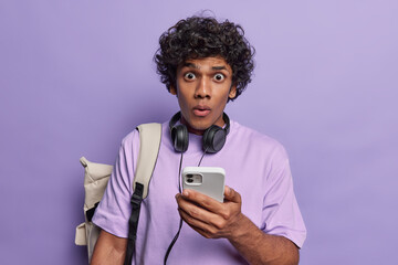Curly haired Hindu man using smartphone writing message chatting with friend surfing internet reacts to something shocking carries rucksack with necessary things dressed in casual t shirt isolated