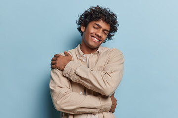 Waist up shot of young Hindu man embraces himself with warm and contented smile creating cozy and reassuring atmosphere captured in moment of self affection and comfort wears brown shirt poses indoor