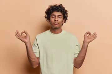 Foto op Canvas Calm young curly haired young Hindu man posing with closed eyes practicing yoga trying to relax finds inner peace dressed in casual basic tshirt isolated over brown background enjoying free time © Wayhome Studio