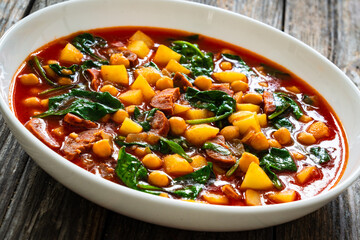 Delicious Spanish stew - chickpeas, spinach, seared chorizo, potatoes, tomatoes and broth on wooden table
