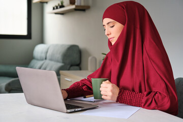 Muslim young woman university student in hijab drinking coffee and using the laptop at home.