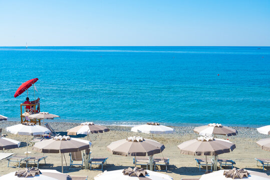 Lifeguard under red umbrella in one of the beaches of Varazze, Liguria, Italy, with umbrella rows and peaceful blue waters. The writings on the red umbrella means "Lifeguard" in several languages.