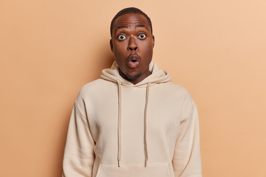Attractive dark skinned man witnessing something truly extraordinary evoking sense of wonder and shared exhilaration stands speechless dressed in casual sweatshirt isolated over brown background