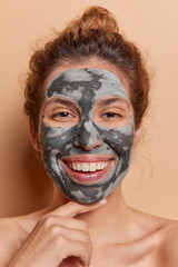 Vertical shot of glad European woman infusing skin with revitalizing minerals and nutrients applies facial clay mask touches chin gently stands shirtless isolated over brown background. Beauty concept