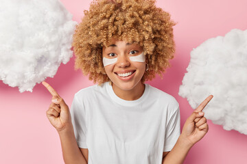 Glad curly haired woman applies beauty patches under eyes for skin rejuvenation points both index fingers overhead smiles pleasantly dressed in casual white t shirt shows something on clouds