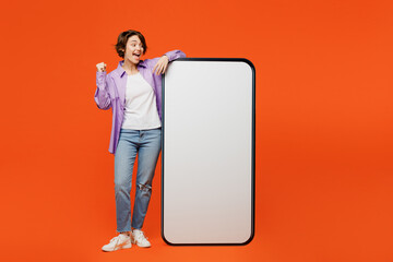 Full body young woman she wear purple shirt white t-shirt casual clothes big huge blank screen mobile cell phone smartphone with area doing winner gesture isolated on plain orange background studio.