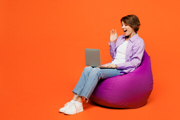 Full body happy young IT woman she wear purple shirt white t-shirt casual clothes sit in bag chair hold use work on laptop pc computer waving hand isolated on plain orange background studio portrait.