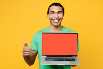 Close up young IT man of African American ethnicity in casual clothes green t-shirt hat work hold use laptop pc computer blank screen workspace area show thumb up isolated on plain yellow background