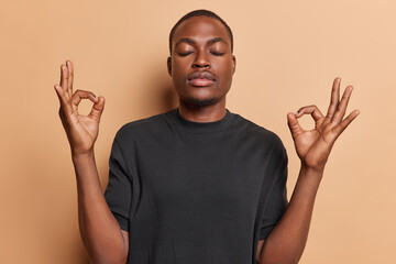 Calm dark skinned man enjoys morning yoga practicing breathing exercises for inner peace and harmony meditates and keeps hands in okay gesture wears black t shirt isolated over brown background