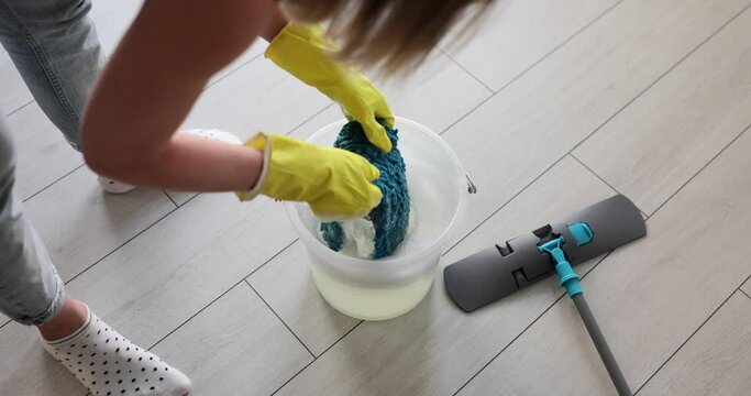 Woman in yellow gloves rinsing mop cloth in bucket of detergent after damp cleaning of house. Housewife cleans parquet floor at home
