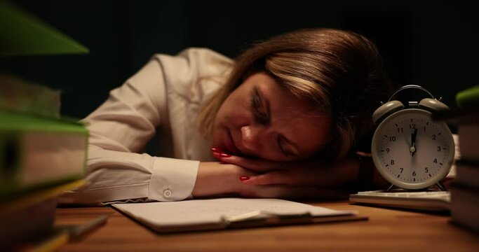 Exhausted female worker lies on hands put on office table resting near folders with documents and vintage alarm clock. Tired woman opens eyes trying to start work