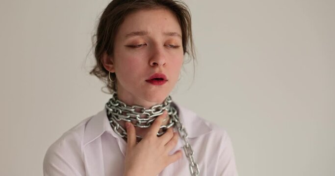 Young woman touches metal chains on throat breathing heavily. Lady with sick throat in white shirt feels discomfort from being chained. Freedom of speech concept
