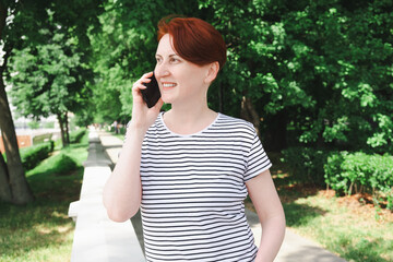 Fototapeta premium Middle aged woman with red hair stands on the embankment of the city park, smiling while talking on the phone. Portrait of a woman in a striped T-shirt.