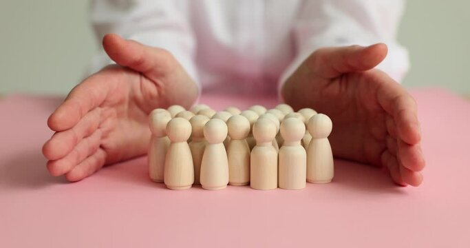 Person covers group of small wooden figurines with hands to protect. Pawns stand on pink background. Teamwork society and community support concept