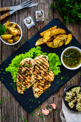 Grilled chicken breasts with Argentinian chimichurri sauce,  baked potatoes and fresh vegetables on wooden table
