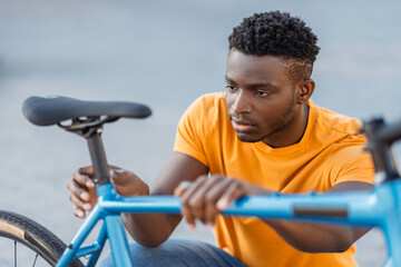 Closeup portrait of attractive Nigerian man checking bicycle seat on street. Concept of cycling - obrazy, fototapety, plakaty