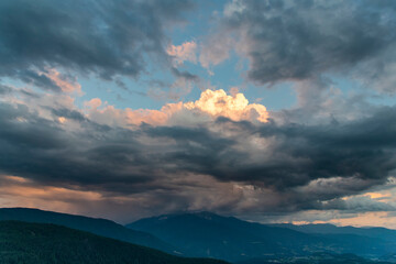 over mountains and valley during sunset with dark thunderclouds hanging over the peak of the mountain with opening in clouds with blue sky and sun coloring cumulus clouds 