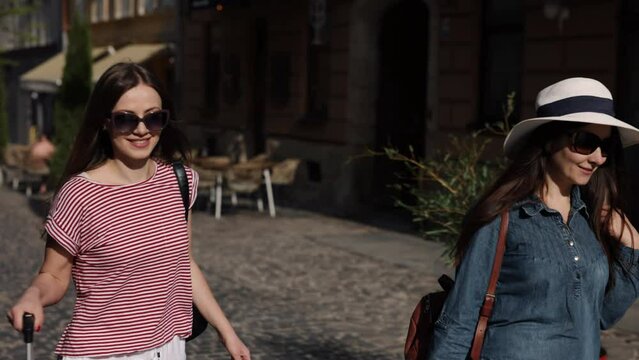 Carrying Suitcase, Summer vacation, Tourist travel. Two good-looking women, clearly tourists, tread down street, their luggage accompanying them, relishing their journey in foreign land.