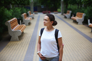 Beautiful multi-ethnic woman in white mockup t-shirt and jeans, with backpack, walking in city park...