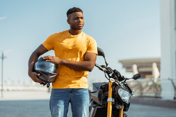 Fototapeta na wymiar Handsome African man holding protective helmet standing near modern motorcycle on street outdoors