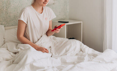 Obraz na płótnie Canvas Young woman with phone lying in comfortable bed. Caucasian smiling 30s girl using smartphone while lying on bed at home, copy space