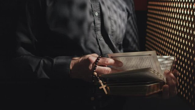 Medium section shot of unrecognizable Catholic priest holding rosary reading Bible or prayer book in confessional