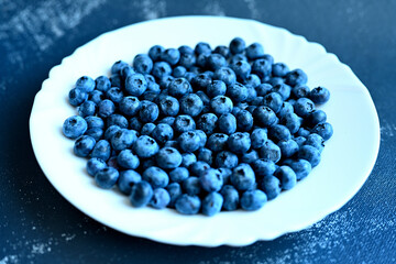 Ripe blueberries on a white plate against a dark gray background, close-up. Organic, nutritious food full of vitamins and minerals.