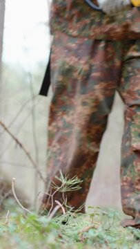 A man dressed in camouflage clothing walks through the woods with a metal detector and a shovel. Search for treasure, coins, metal. Vertical video.
