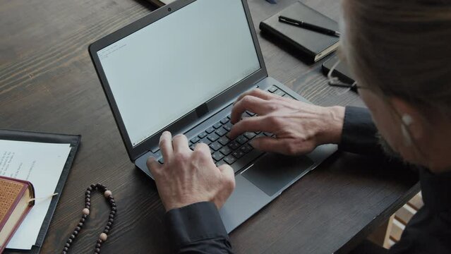 High angle view of unrecognizable mature Catholic pator sitting at wooden table writing essay on laptop