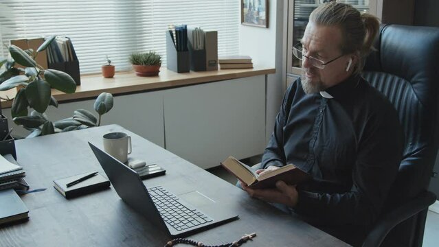 High angle shot of mature Catholic pastor sitting at desk in office checking something in book during online meeting with colleagues