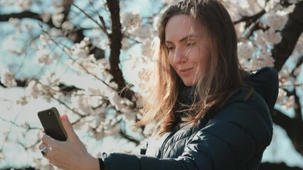 Girl making selfie with white sakura flowers in Tokyo, Japan. Woman enjoy spring blossom happy smiling looking to the camera. Sunny spring outdoors activity. Spring blossom of sakura tree in city park