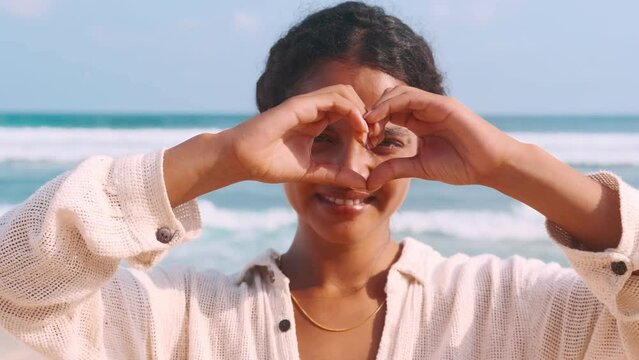 Young beautiful Indian woman teenager smiling looks at camera and shows heart symbol to recommend you go on vacation to south and visit exotic country stands on beach near sea waves.