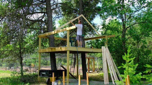 Man checking roof style template while working on tree house and zip line starting platform. There are two large trees going through platform and tree house.