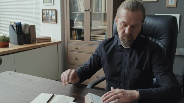 Modern Mature Caucasian priest sitting at desk in his office reading book and writing down quotes in his notebook