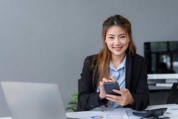 Portrait of a smiling asian businesswoman using mobile phone in the office.  Young Asian businesswoman with paperwork at workplace using smartphone in a modern office.