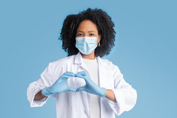 Glad young black woman doctor in white coat, protective mask and gloves shows heart sign with hand